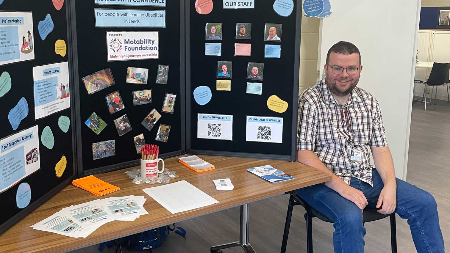 A photo of Robert from our team at our Open Day. He is sat next to an information stall for our Travel with Confidence project.